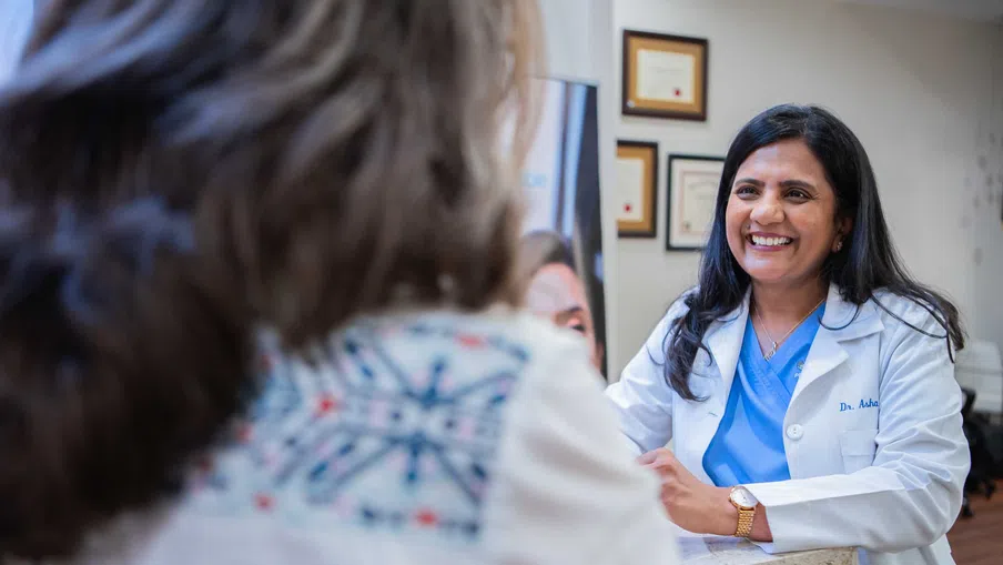 Smiling female doctor in a white coat consulting with a patient across a reception desk in a medical office.