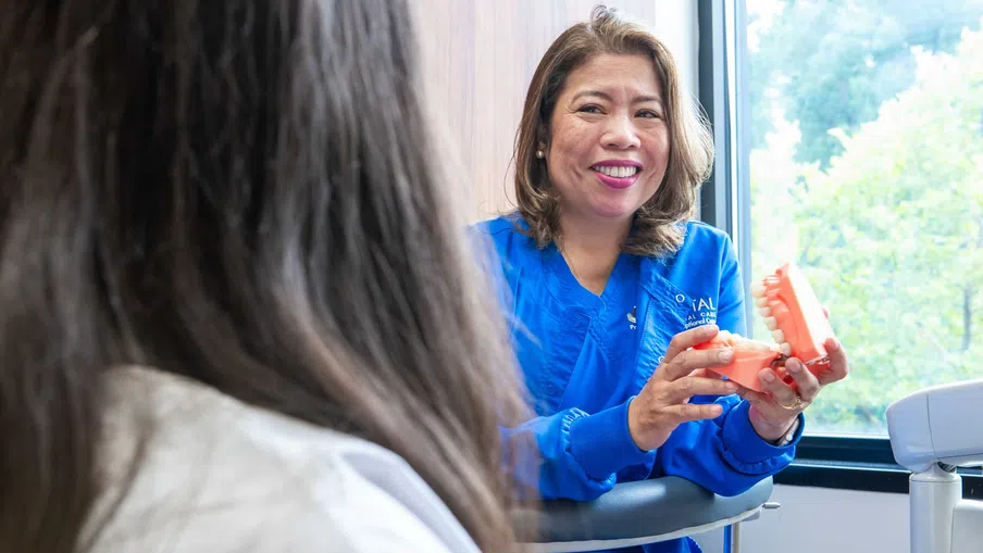 Female dentist in blue scrubs explaining dental care using a tooth model to a patient in a clinic.