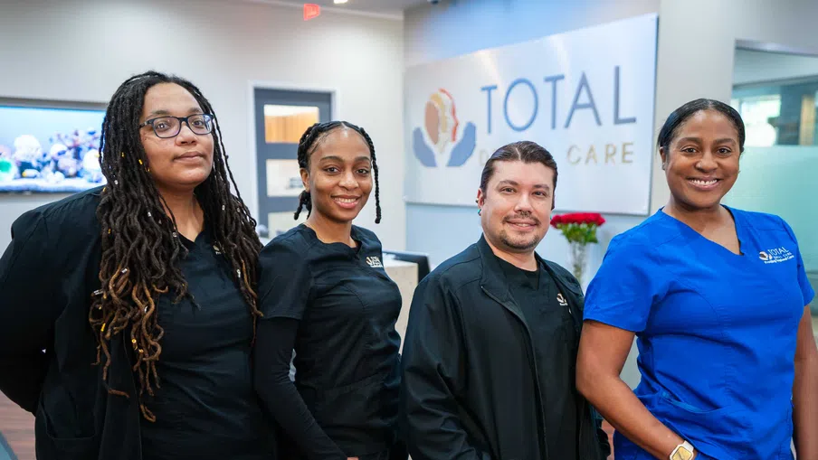 Four healthcare professionals in black and blue scrubs smiling inside a Total Care medical office lobby.
