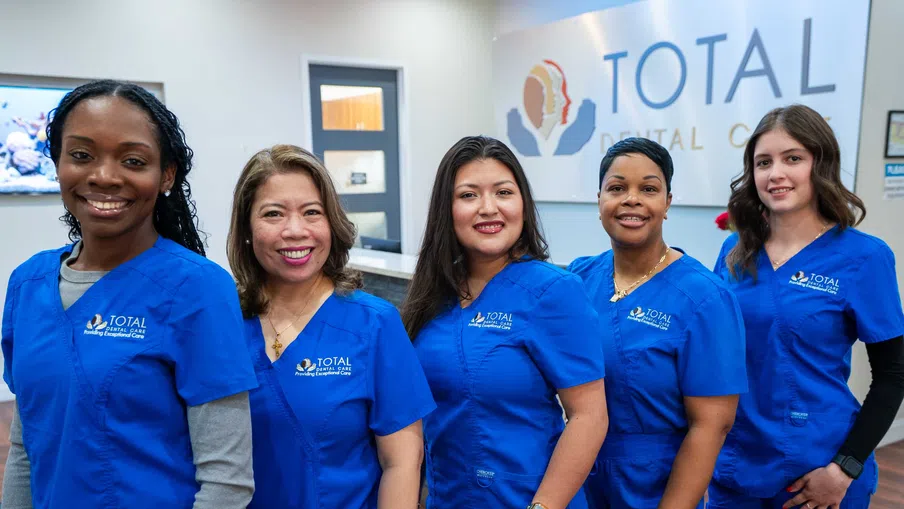 Smiling dental staff in blue uniforms standing in front of Total Dental Care clinic reception desk.