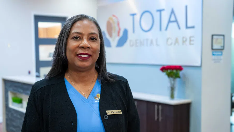 Smiling female dentist in blue scrubs standing in Total Dental Care clinic with flowers and reception desk in background