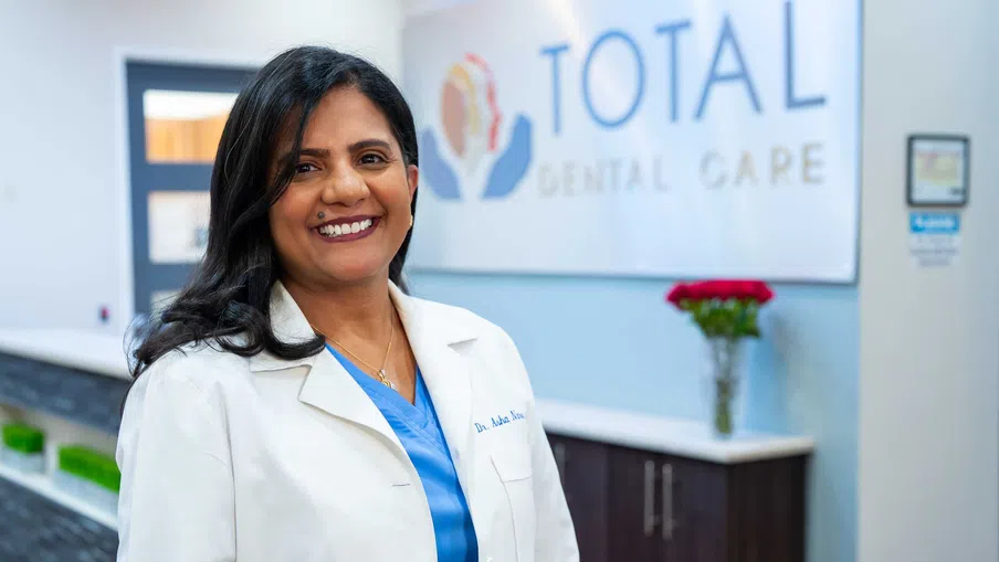 Smiling female dentist in white coat standing inside Total Dental Care clinic with sign in background
