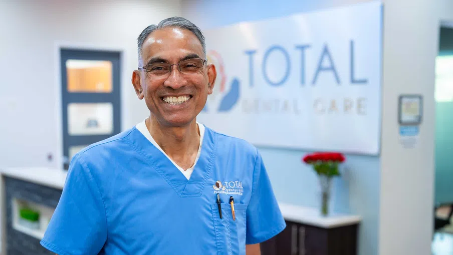 Smiling male dentist in blue scrubs standing inside a modern dental clinic with Total Dental Care sign in background