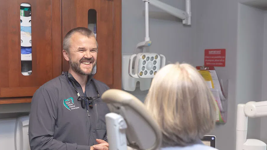 Smiling male dentist talking to a seated female patient in a modern dental clinic room.