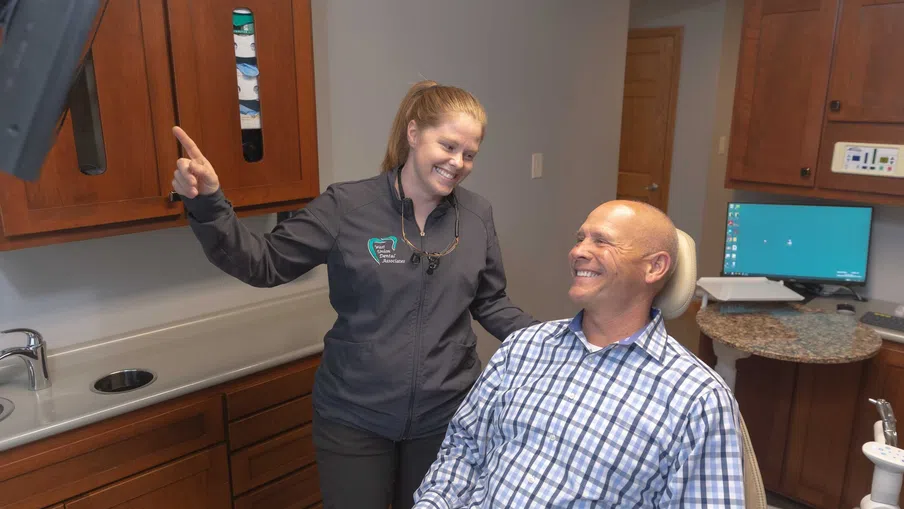 Dentist smiling and explaining treatment to male patient in dental clinic exam room.