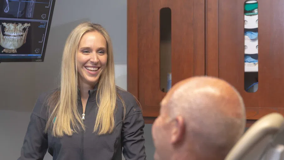 Smiling female dentist talking to a male patient in a dental office with a 3D dental X-ray on screen.