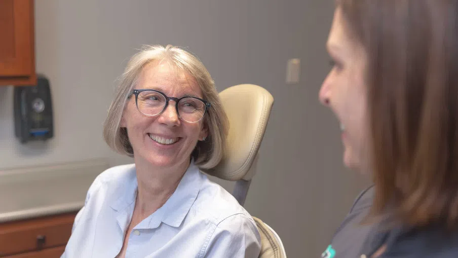 Smiling senior woman wearing glasses sitting in a dental chair talking to a female dentist.