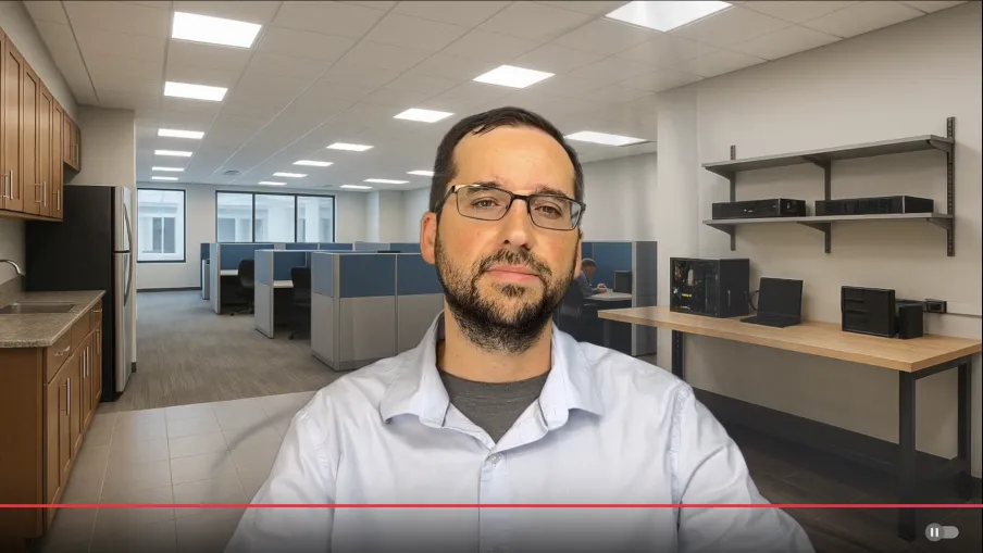 Man with glasses and beard sitting in a modern office with cubicles and a kitchen area