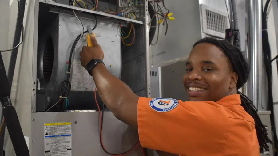 Estes Services Technician in orange uniform smiling while repairing HVAC system with tools and exposed wiring.