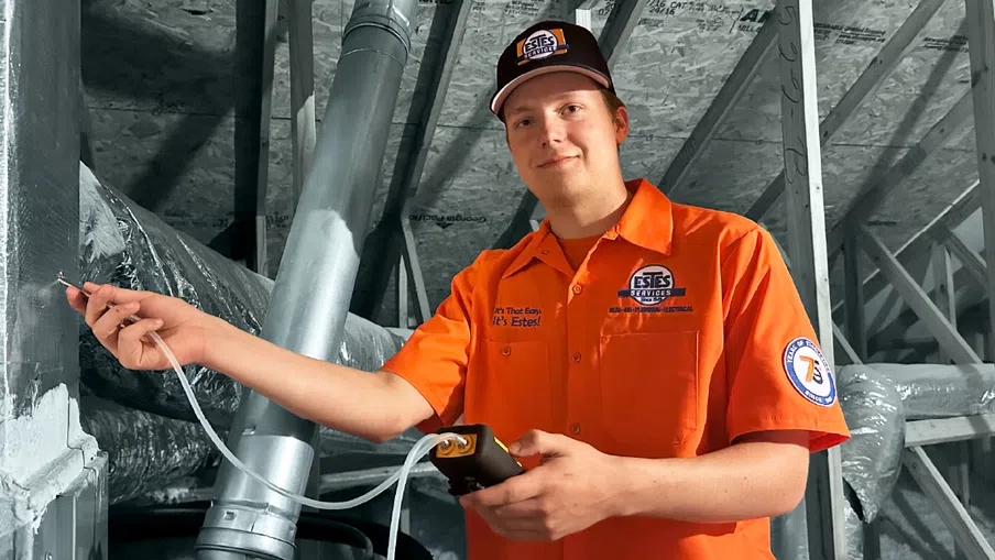 Technician in an orange Estes Services uniform testing HVAC ductwork with digital equipment in an attic space.