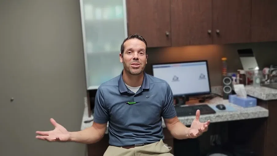 Man in blue polo shirt sits in an office with computer and cabinets, gesturing with open hands.