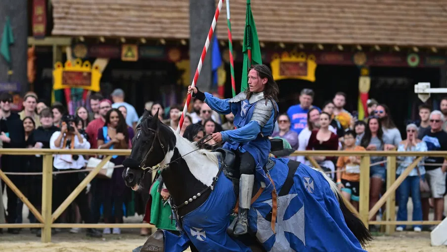 Knight in blue armor rides a black and white horse holding a lance during a medieval jousting event with spectators.