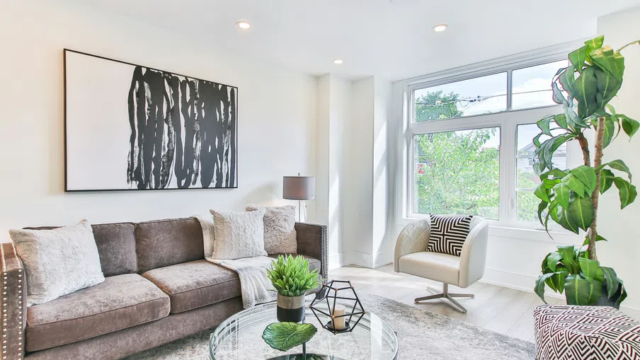 Bright living room with brown sofa, glass coffee table, large window, modern armchair, and indoor plants.