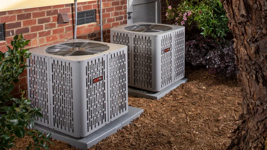 Two gray outdoor air conditioning units installed beside a brick house with mulch and plants around.