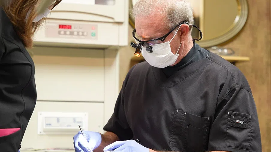 Dentist wearing magnifying glasses and gloves performing dental procedure on patient in clinic.