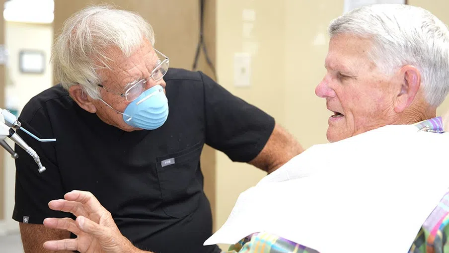 Dentist wearing mask talks to elderly male patient during dental checkup in clinic.