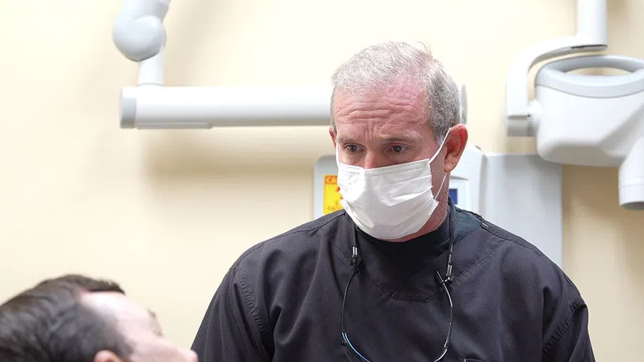 Dentist wearing a mask and protective clothing examining a male patient in a dental clinic.