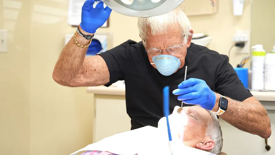 Dentist wearing mask and gloves examining elderly patient's teeth under dental light in clinic