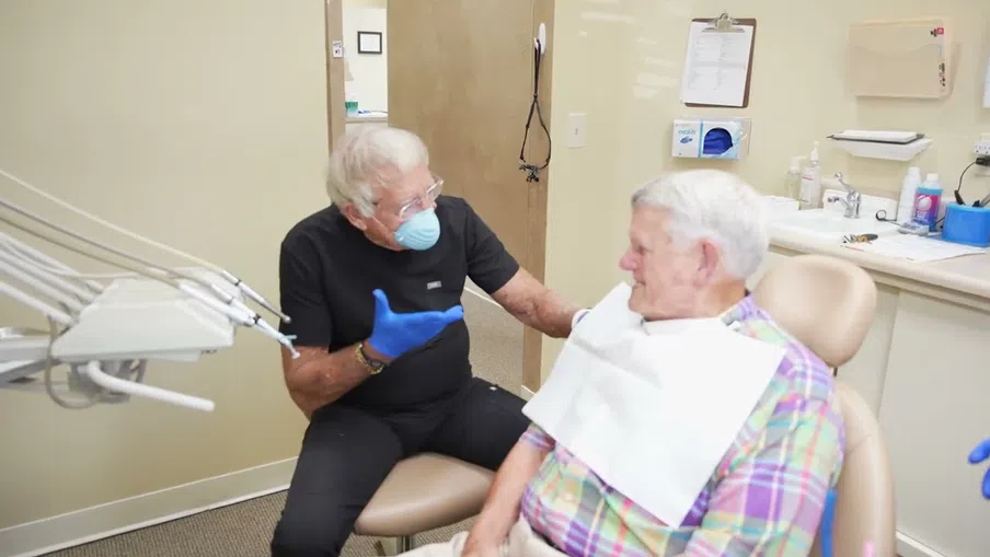 Dentist wearing mask and gloves talking to elderly male patient in dental clinic chair