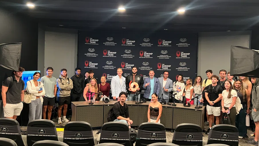 Group photo in conference room with diverse attendees posing behind desk and Shrimp Tank branded backdrop.
