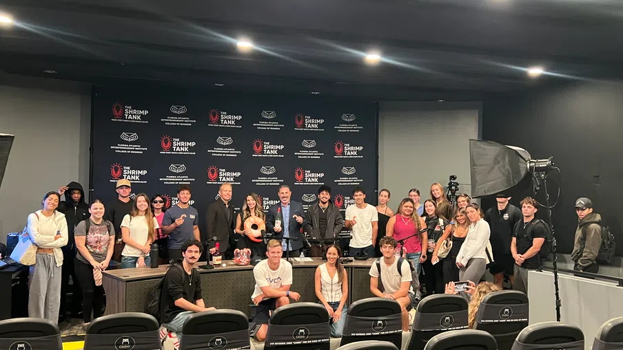 Group photo of diverse people standing and sitting in a studio with The Shrimp Tank branded backdrop and lighting.