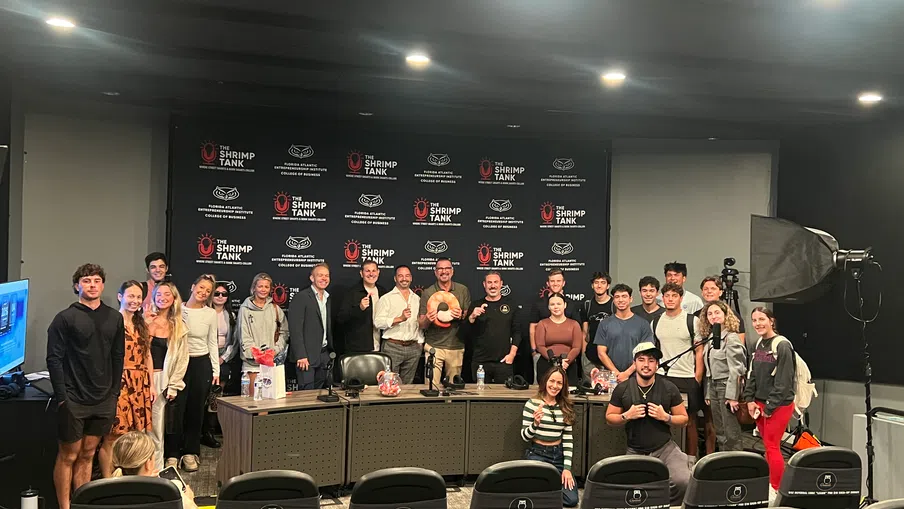 Group of people posing in a conference room with a 'The Shrimp Tank' backdrop, with overlaid text logos