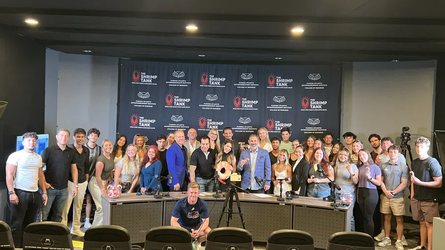 Large group of people posing in front of The Shrimp Tank backdrop with conference chairs in the foreground