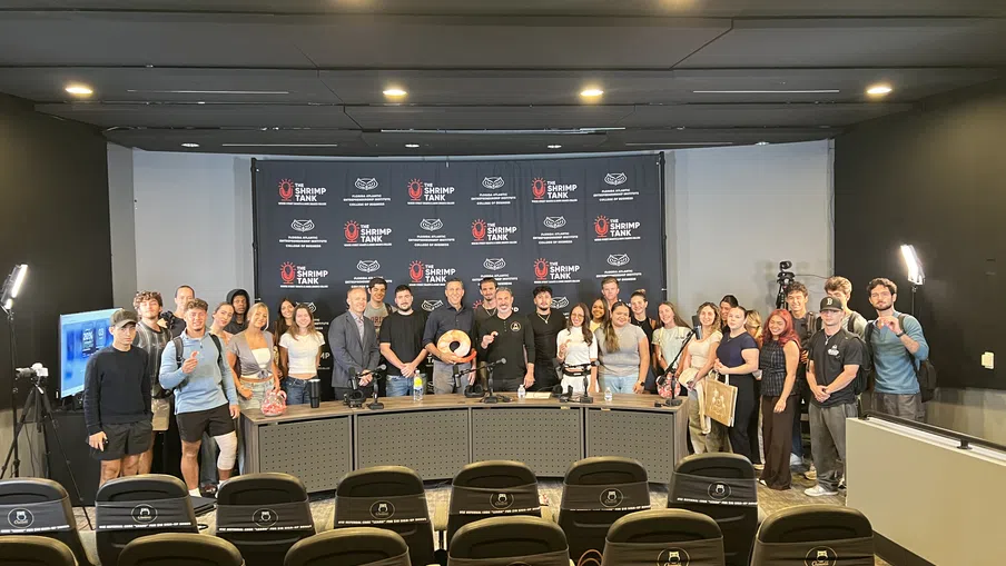 Group photo of diverse people standing behind a table with The Shrimp Tank backdrop in a conference room.