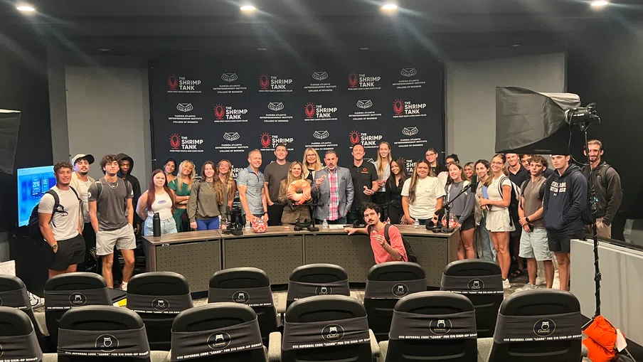 Group photo of young adults on a Shrimp Tank studio set with empty audience seats and studio lighting visible