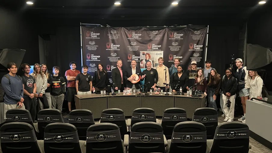Group of people posing behind a table in a studio with The Shrimp Tank backdrop and empty audience chairs in front.