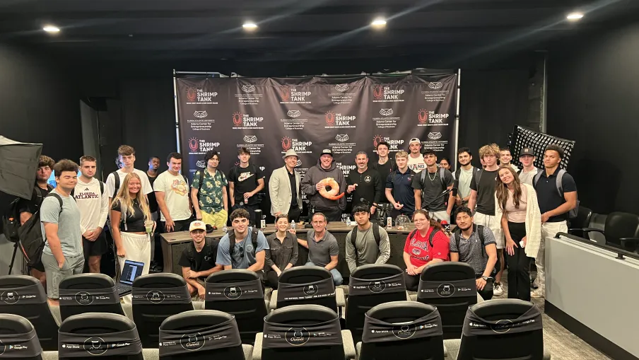 Group of young adults and instructors posing in front of a backdrop with Shrimp Tank logos in a modern conference room.