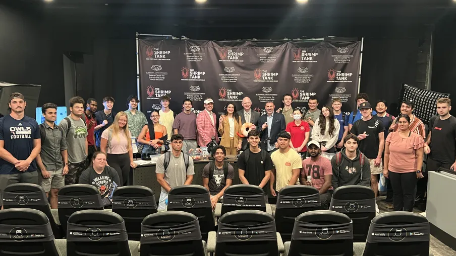 Large group of diverse young adults and instructors posing indoors in front of a black backdrop with Shrimp Tank branding.