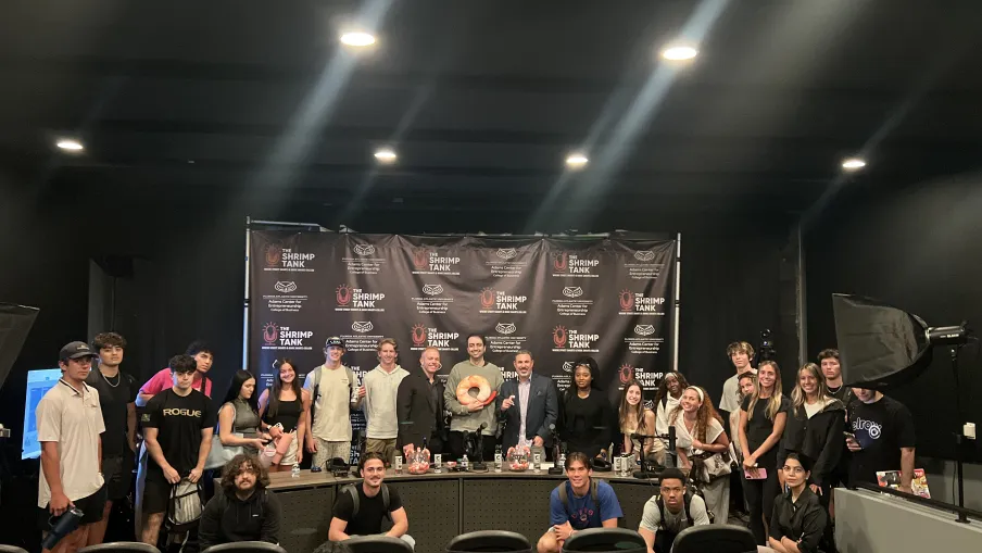 Group of young adults posing in a studio with The Shrimp Tank backdrop and workstations under bright lights.