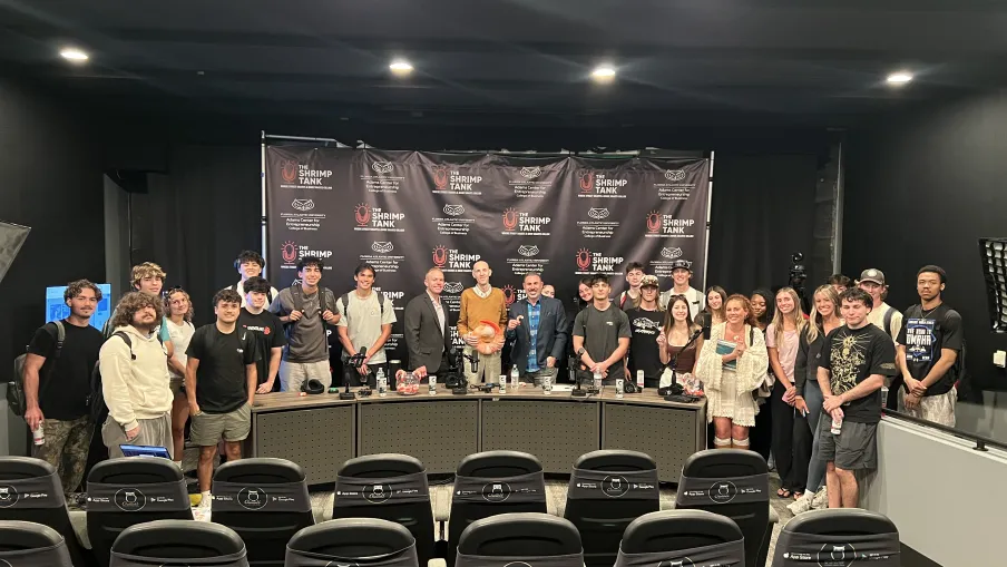 Group photo of diverse people standing behind a panel table in a conference room with The Shrimp Tank backdrop.