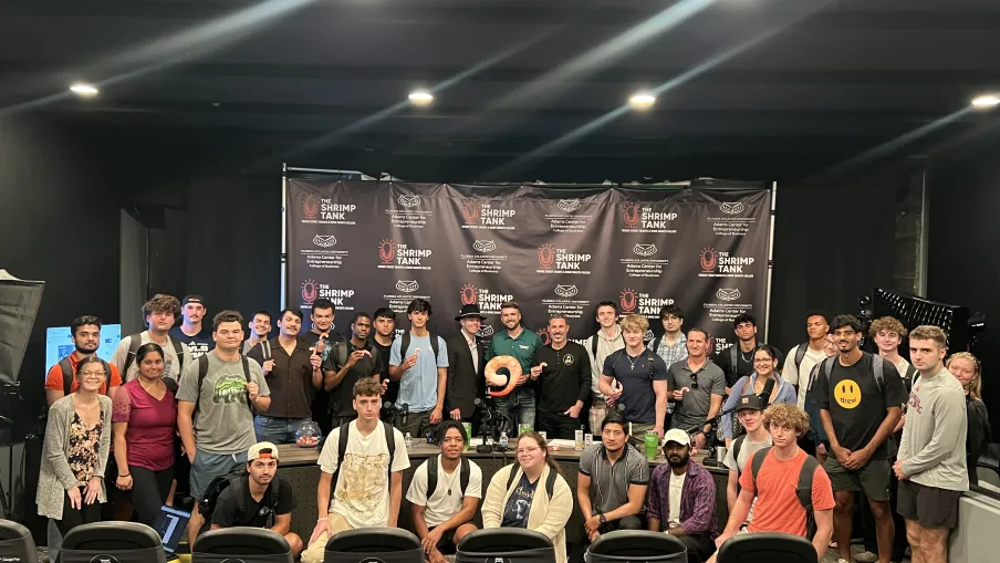 Large group of diverse young adults and instructors posing indoors in front of a black backdrop with Shrimp Tank branding.