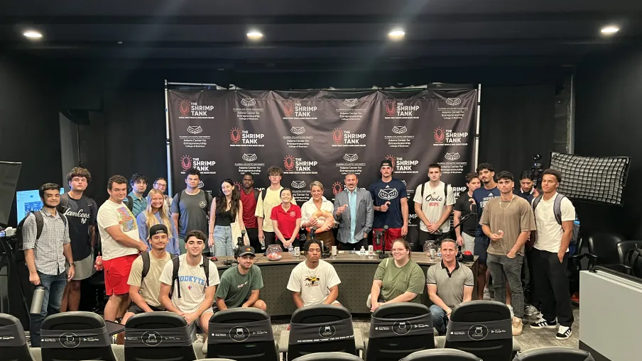 Group photo of diverse young adults and instructors posing in front of a The Shrimp Tank branded backdrop in a studio.