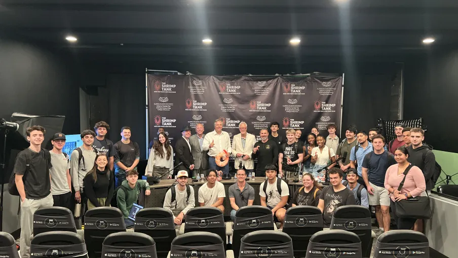 Group of young adults and professionals posing for a photo in a studio with a Shrimp Tank backdrop and empty theater seats in front.