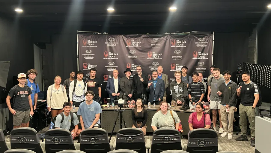 Group of young adults and instructors posing in front of a backdrop with Shrimp Tank logos in a modern conference room.