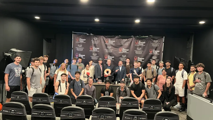 Large group of young adults and professionals pose in front of a Shrimp Tank branded backdrop in studio setting.