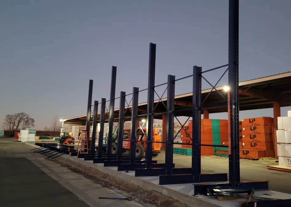 Steel framework under construction in a lumber yard during twilight with stacked wood panels in the background