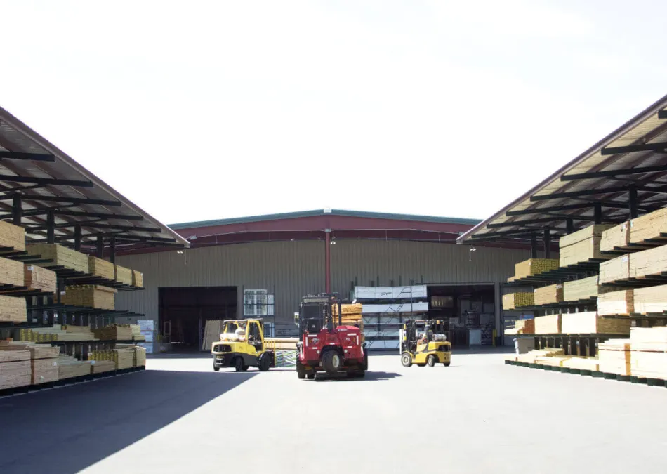 Outdoor lumber yard with stacked wooden planks and forklifts operating in front of a warehouse under clear sky.