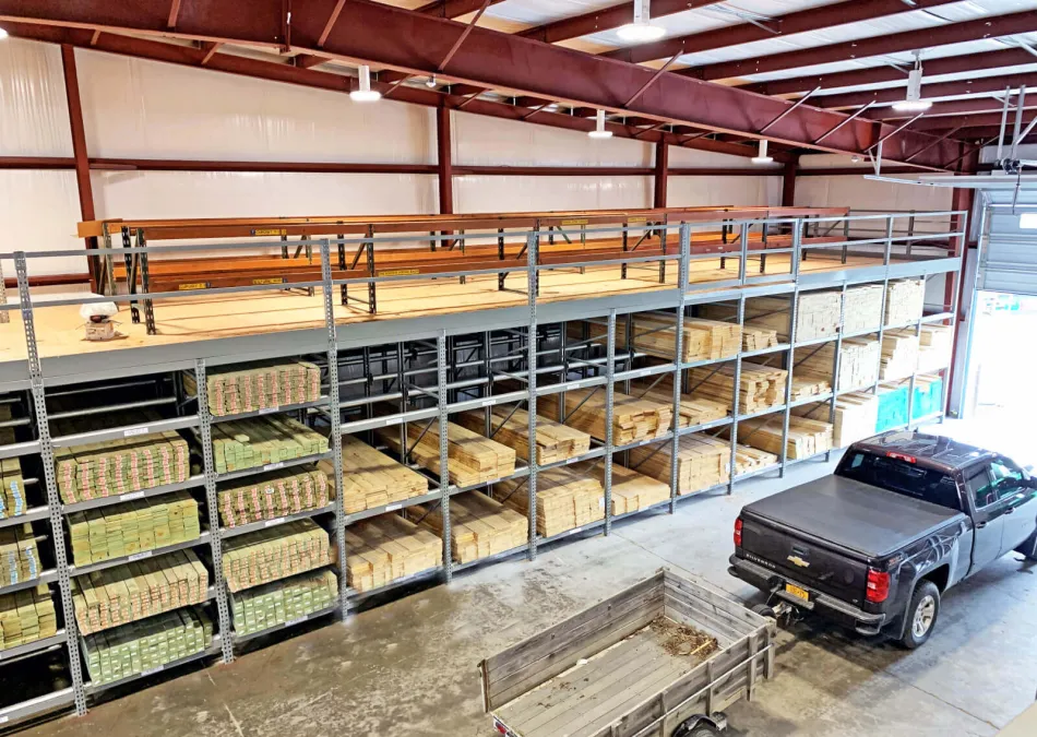 Warehouse interior with metal shelving stocked with lumber and a black pickup truck with trailer inside