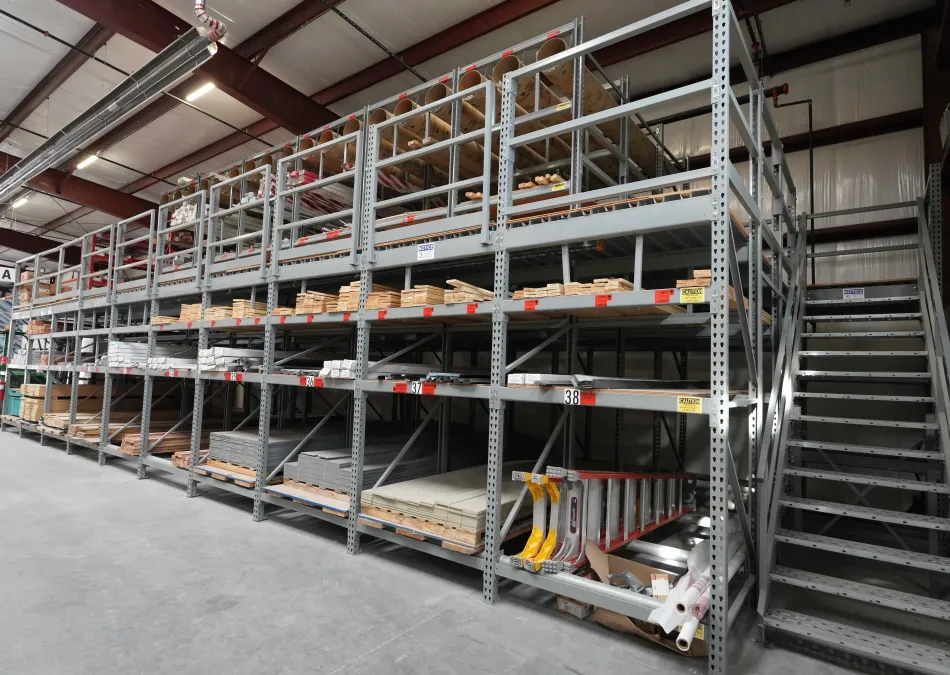 Metal shelving units in a large warehouse storing wood and construction materials with a metal staircase beside them.