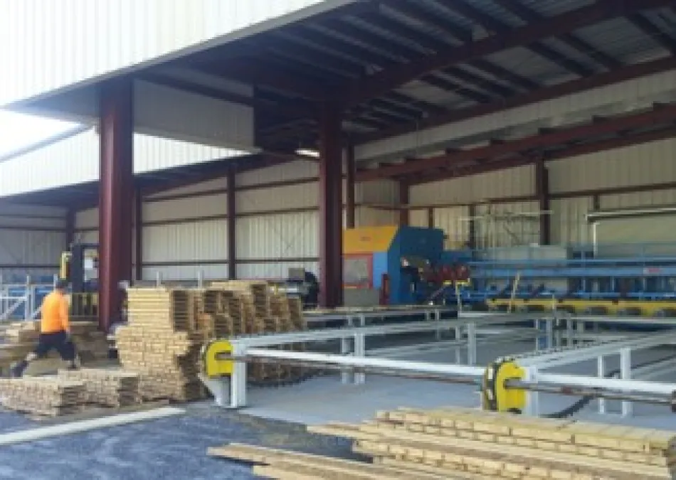 Worker stacking wooden planks inside a spacious industrial warehouse with metal machinery and open roof structure
