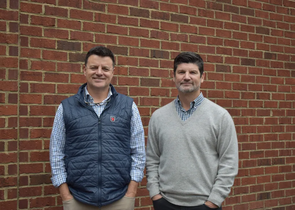 Two men standing against a red brick wall, one wearing a blue vest and the other a gray sweater, both with hands in pockets.