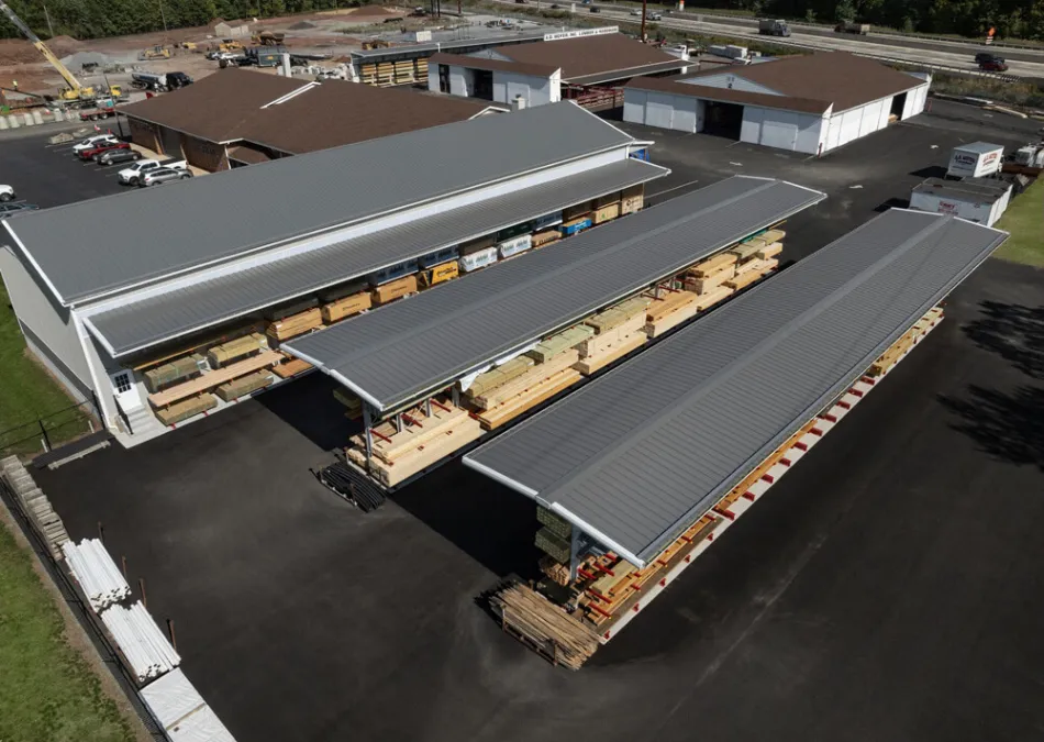 Aerial view of a lumber yard with stacks of wood stored under metal-roofed shelters and adjacent buildings.