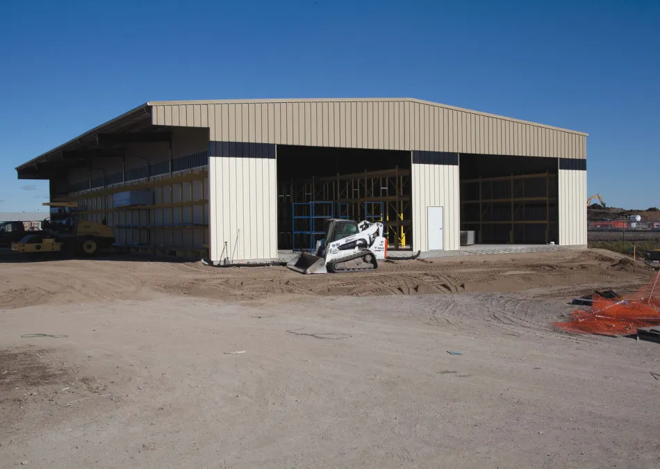 Large beige metal warehouse under construction with dirt ground and small construction vehicle in front