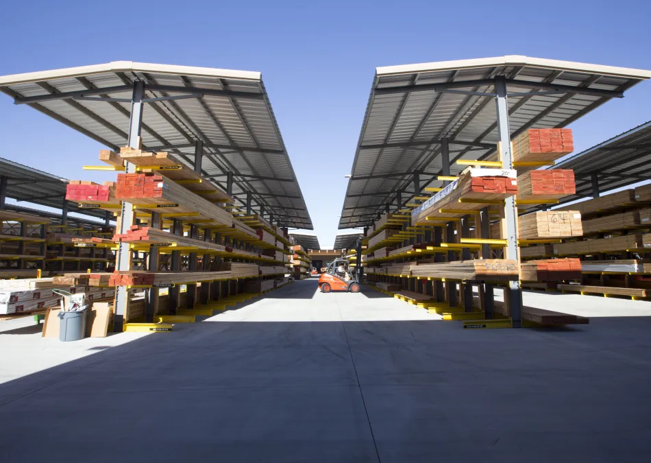 Outdoor lumber yard with organized stacks of wood under metal canopies and a forklift in the center aisle.