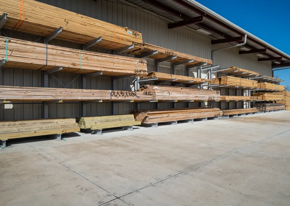 Stacks of wooden lumber neatly arranged on metal racks outside a building under a clear blue sky.