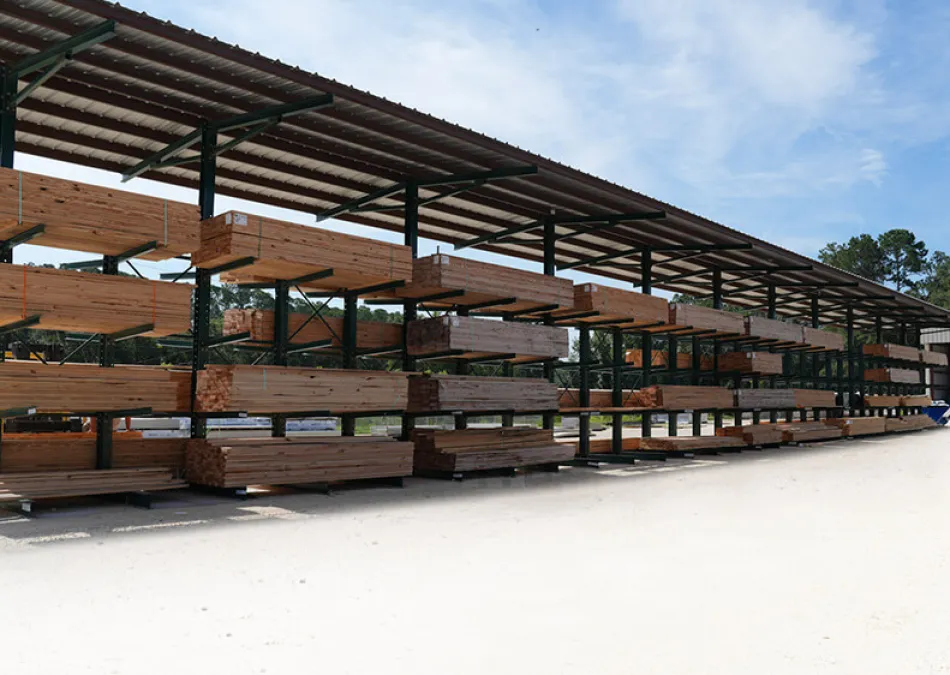 Outdoor lumber storage racks filled with stacked wooden planks under a metal roof on a clear day.